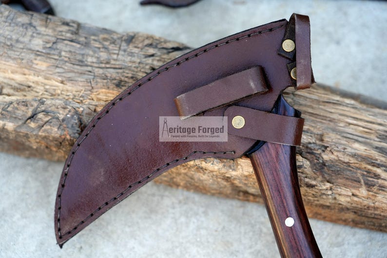 Op de afbeelding: Een bruine leren schede met een bruine houten handvat en een metalen mes. De schede heeft een riem en een metalen klinknagel. De tekst "Heritage Forged" is op de schede gedrukt.