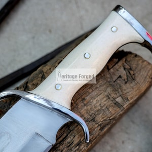 Op de afbeelding: Close-up van een mes met een witte handgreep en een zilveren lemmet. De handgreep heeft een decoratief ontwerp en de woorden "Heritage Forged" zijn op het lemmet gedrukt. Het mes ligt op een stuk hout.