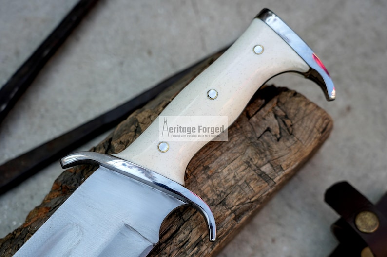Op de afbeelding: Close-up van een mes met een witte handgreep en een zilveren lemmet. De handgreep heeft een decoratief ontwerp en de woorden "Heritage Forged" zijn op het lemmet gedrukt. Het mes ligt op een stuk hout.