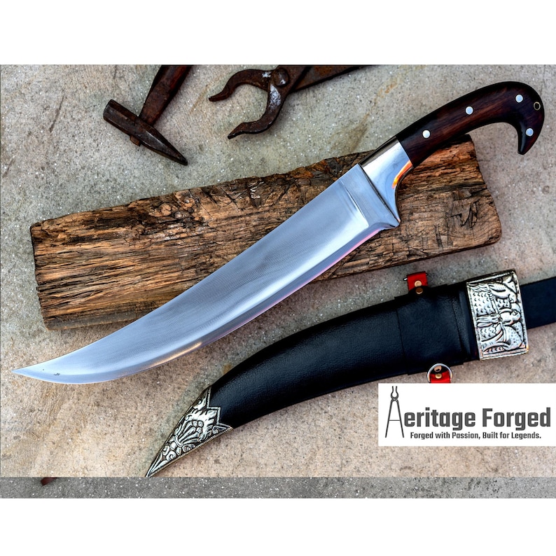May include: A curved, silver-colored knife with a dark wood handle and a black leather sheath with silver accents. The knife is resting on a wooden surface. The text "Heritage Forged" is visible in the lower right corner of the image. "Forged with Passion, Built for Legends."