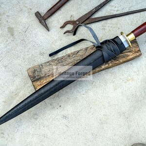 May include: A black dagger with a wooden handle and a leather sheath rests on a wooden plank. The handle is dark red with a gold-colored metal guard. Black leather straps wrap around the sheath. Blacksmith tools, including a hammer and pliers, are in the background.