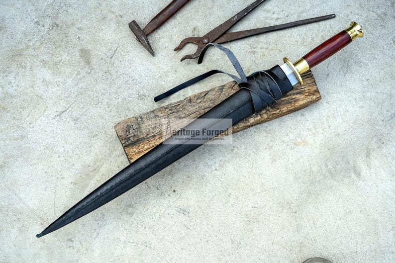 May include: A black dagger with a wooden handle and a leather sheath rests on a wooden plank. The handle is dark red with a gold-colored metal guard. Black leather straps wrap around the sheath. Blacksmith tools, including a hammer and pliers, are in the background.