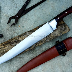 May include: A long, silver knife with a brown wooden handle and a leather sheath. The knife is resting on a piece of wood with the text "Heritage Forged" visible. The knife is surrounded by other tools, including a hammer and pliers.