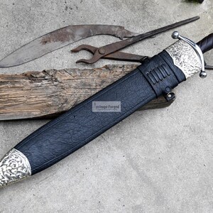 May include: A silver and black sword with a leather scabbard. The sword has a decorative handle and a pointed blade. The scabbard is black leather with a silver tip.