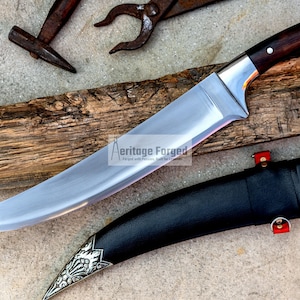 May include: A curved, silver-colored knife with a dark wood handle and a black leather sheath. The knife has a decorative silver design on the sheath. The text "Heritage Forged" is visible on the blade.