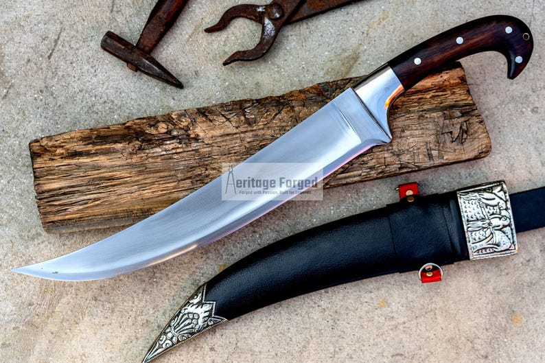 May include: A curved, silver-colored knife with a dark wood handle and a black leather sheath. The knife has a decorative silver design on the sheath. The text "Heritage Forged" is visible on the blade.