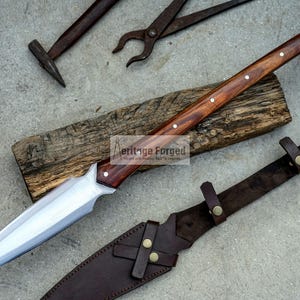 K&ouml;nnte beinhalten: Eine lange, scharfe, silberne Klinge mit einem Holzgriff und Messingakzenten. Die Klinge liegt auf einem St&uuml;ck Holz mit dem aufgedruckten Text "Heritage Forged". Eine braune Lederscheide mit Messingakzenten ist ebenfalls abgebildet.