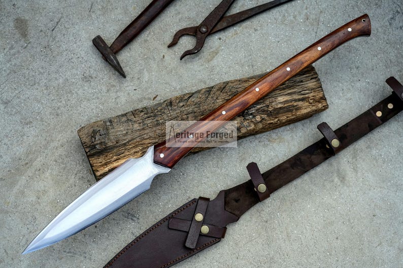 K&ouml;nnte beinhalten: Eine lange, scharfe, silberne Klinge mit einem Holzgriff und Messingakzenten. Die Klinge liegt auf einem St&uuml;ck Holz mit dem aufgedruckten Text "Heritage Forged". Eine braune Lederscheide mit Messingakzenten ist ebenfalls abgebildet.
