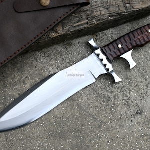 Puede incluir: Un cuchillo de caza grande, de color plateado, con un mango de madera marr&oacute;n. El cuchillo tiene un filo dentado y una punta afilada. El mango tiene un patr&oacute;n texturizado y una guarda de metal. El cuchillo est&aacute; sobre una superficie gris.