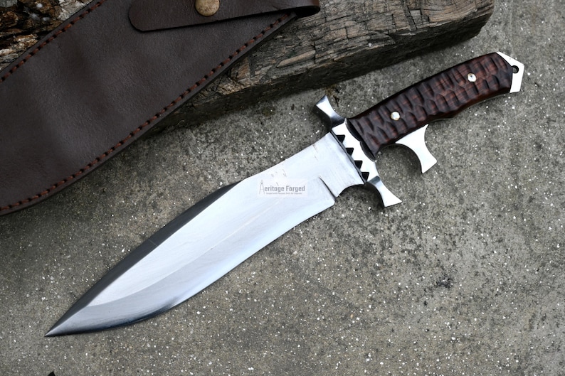 Puede incluir: Un cuchillo de caza grande, de color plateado, con un mango de madera marr&oacute;n. El cuchillo tiene un filo dentado y una punta afilada. El mango tiene un patr&oacute;n texturizado y una guarda de metal. El cuchillo est&aacute; sobre una superficie gris.