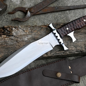 Puede incluir: Un cuchillo grande de color plateado con un mango de madera marr&oacute;n. El cuchillo tiene un filo dentado y una punta afilada. El cuchillo est&aacute; apoyado sobre una superficie de madera con una funda de cuero marr&oacute;n.