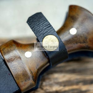 May include: Close-up of a knife handle with a dark brown wood finish and a black leather sheath. The handle has a brass accent with the text "Heritage Forged" on it.