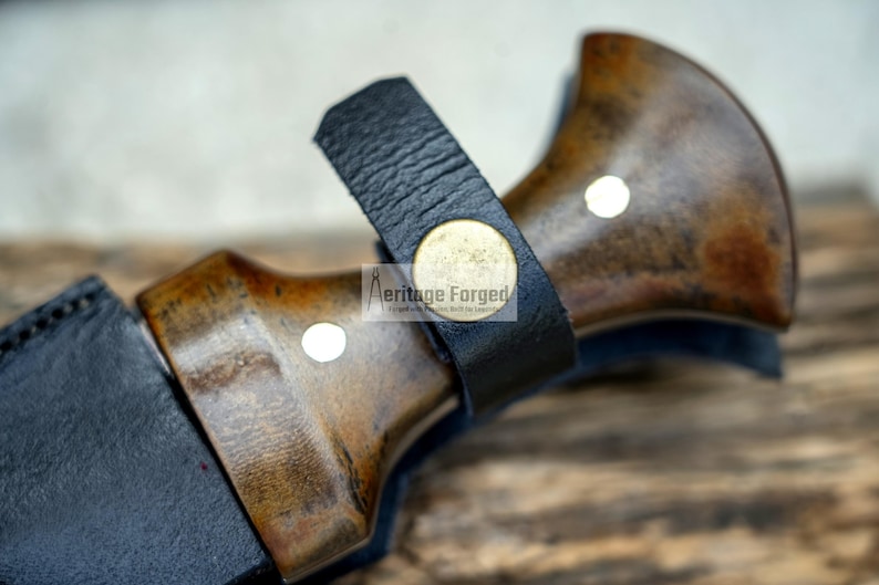 May include: Close-up of a knife handle with a dark brown wood finish and a black leather sheath. The handle has a brass accent with the text "Heritage Forged" on it.