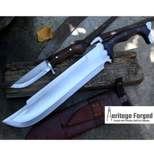 May include: A large, silver-colored knife with a brown wooden handle. The knife has a curved blade and a sharp point. The knife is resting on a wooden surface, and there is a smaller knife with a brown wooden handle in the background. The text "Heritage Forged, Forged with Passion, Built for Legends." is visible in the lower right corner of the image.