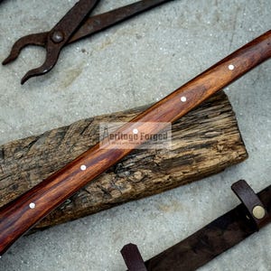 K&ouml;nnte beinhalten: Eine Axt mit einem Holzstiel, einer Klinge aus Metall und einer braunen Lederscheide. Der Stiel hat drei silberne Nieten. Der Text "Heritage Forged" ist auf dem Stiel sichtbar.