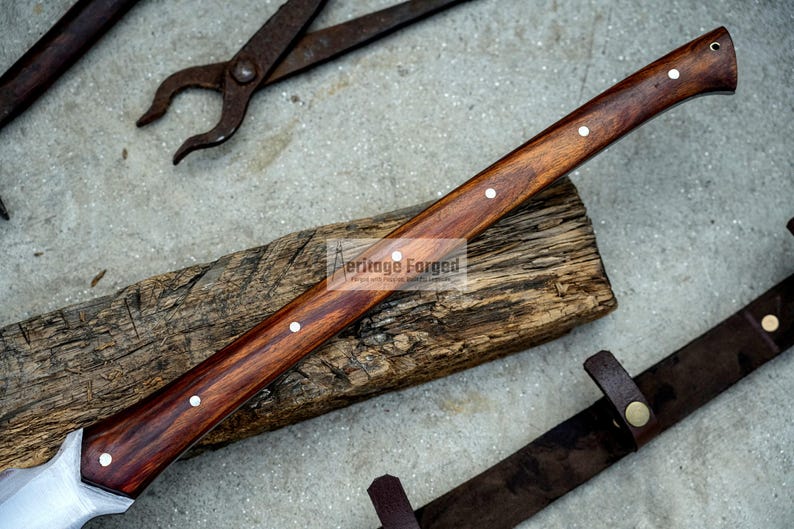 K&ouml;nnte beinhalten: Eine Axt mit einem Holzstiel, einer Klinge aus Metall und einer braunen Lederscheide. Der Stiel hat drei silberne Nieten. Der Text "Heritage Forged" ist auf dem Stiel sichtbar.