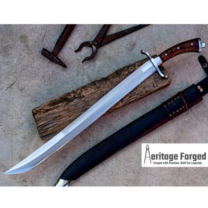 May include: A silver sword with a wooden handle and a black leather scabbard. The sword is lying on a wooden surface with a hammer and pliers in the background. The text "Heritage Forged" is visible in the lower right corner of the image.