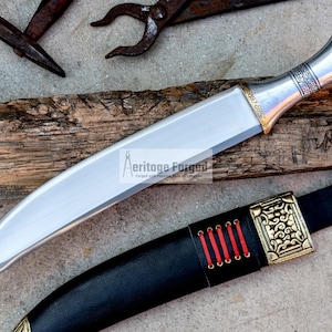 May include: A silver and gold dagger with a curved blade and a decorative handle. The dagger is resting on a piece of wood and has a black leather sheath with gold accents.