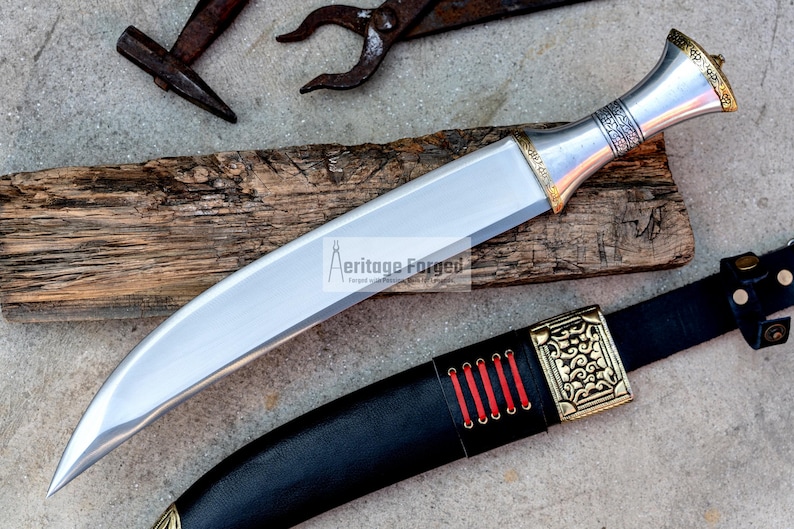 May include: A silver and gold dagger with a curved blade and a decorative handle. The dagger is resting on a piece of wood and has a black leather sheath with gold accents.