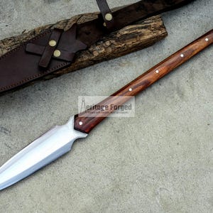 K&ouml;nnte beinhalten: Eine lange, silberne Klinge mit einem Holzgriff. Der Griff ist dunkelbraun mit einem hellbraunen Streifen, der in der Mitte verl&auml;uft. Die Klinge ist scharf und spitz. Das Messer befindet sich in einer Lederscheide mit einer Metallschnalle. Der Text "Heritage Forged" ist auf der Klinge sichtbar.