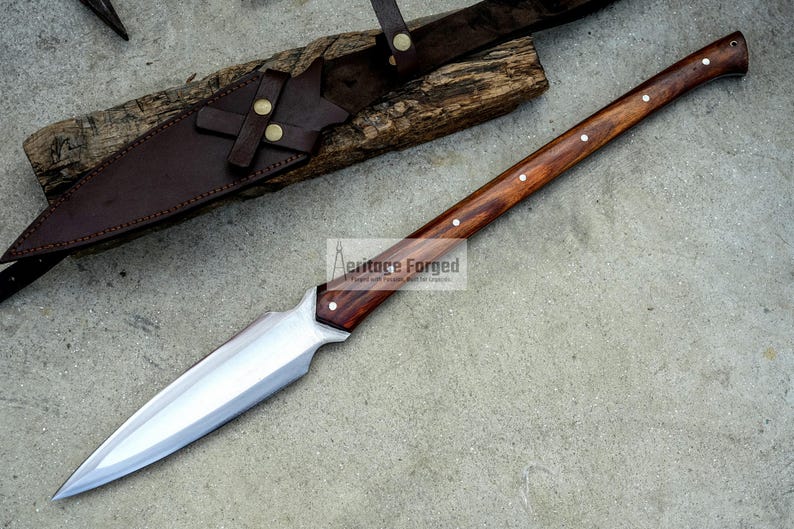 K&ouml;nnte beinhalten: Eine lange, silberne Klinge mit einem Holzgriff. Der Griff ist dunkelbraun mit einem hellbraunen Streifen, der in der Mitte verl&auml;uft. Die Klinge ist scharf und spitz. Das Messer befindet sich in einer Lederscheide mit einer Metallschnalle. Der Text "Heritage Forged" ist auf der Klinge sichtbar.