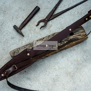 K&ouml;nnte beinhalten: Ein braunes Lederscheide mit einem Riemen und Messingakzenten. Die Scheide liegt auf einem St&uuml;ck Holz mit einem Hammer und einer Zange im Hintergrund. Der Text "Heritage Forged" ist auf dem Holz sichtbar.