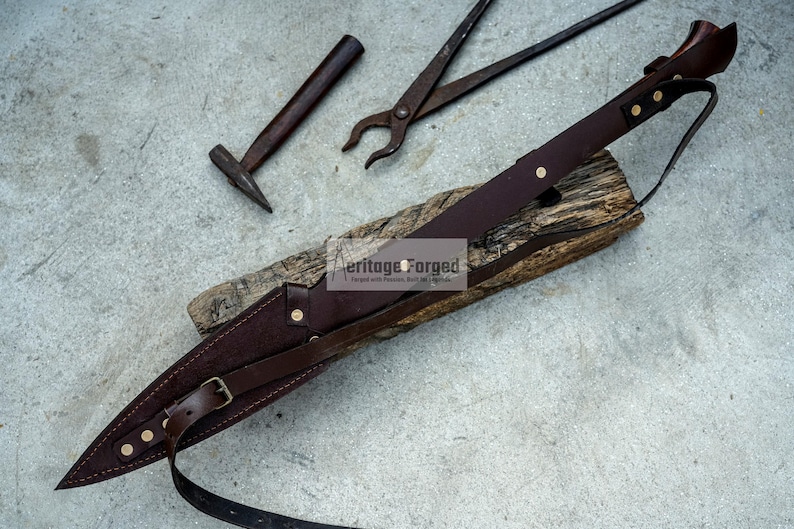 K&ouml;nnte beinhalten: Ein braunes Lederscheide mit einem Riemen und Messingakzenten. Die Scheide liegt auf einem St&uuml;ck Holz mit einem Hammer und einer Zange im Hintergrund. Der Text "Heritage Forged" ist auf dem Holz sichtbar.