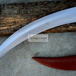 May include: A curved, silver-colored knife with a wooden handle. The knife is resting on a piece of wood with the text "Heritage Forged" printed on it.