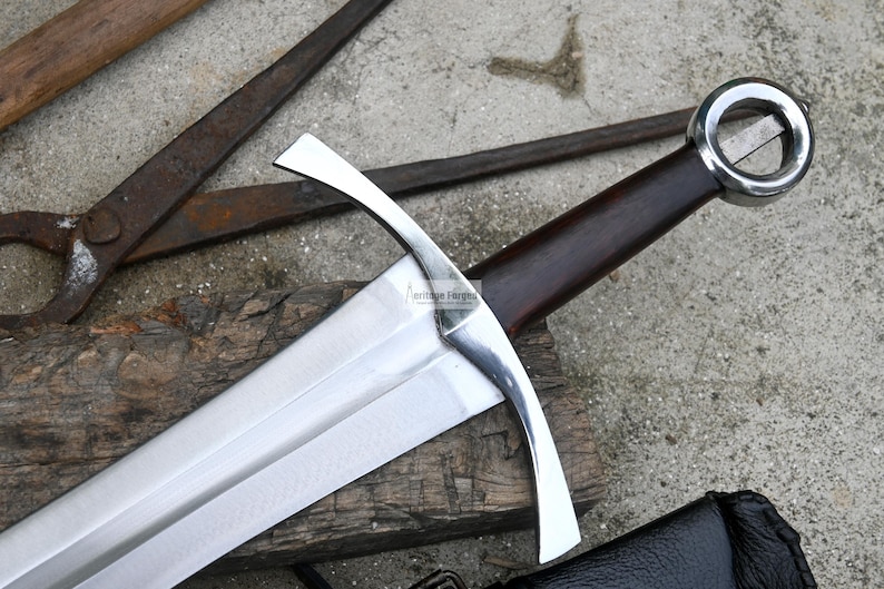Op de afbeelding: Een zilveren middeleeuws zwaard met een bruine houten handvat en een ronde bescherming. Het zwaard ligt op een houten oppervlak.