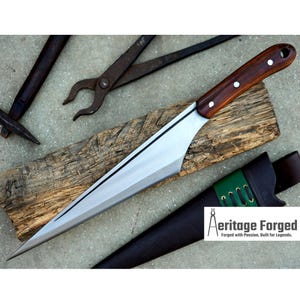 May include: A long, sharp, silver knife with a wooden handle. The knife is resting on a piece of wood and has a leather sheath with a green accent. The text "Heritage Forged" is visible in the lower right corner of the image.  Forged with Passion. Built for Legends.