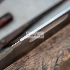Op de afbeelding: Close-up van de rand van een zilveren mesblad met een zwart handvat. Het mes ligt op een stuk hout. De tekst "Heritage Forged" is zichtbaar op het blad.