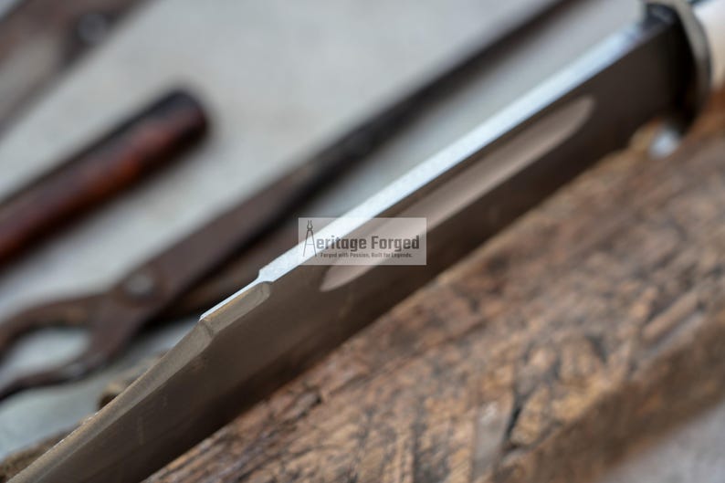 Op de afbeelding: Close-up van de rand van een zilveren mesblad met een zwart handvat. Het mes ligt op een stuk hout. De tekst "Heritage Forged" is zichtbaar op het blad.