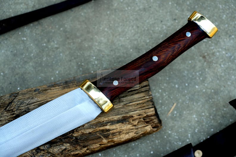 May include: A close-up of a knife with a wooden handle and a brass pommel. The handle is dark brown with a wood grain pattern and two brass rivets. The blade is silver and has a slight curve. The knife is resting on a piece of wood.