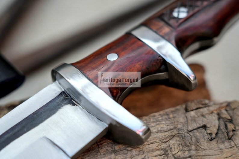 May include: Close-up of a knife with a wooden handle. The handle is a dark reddish brown with a silver metal band around it. The blade is silver and has a slight curve to it. The text "Heritage Forged" is visible on the handle.