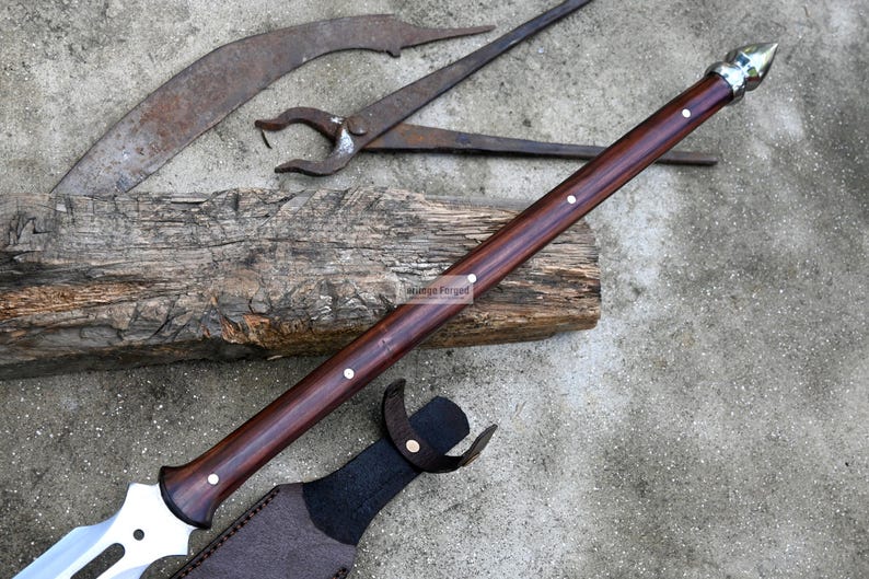 May include: A brown wooden handled spear with a silver metal tip and a leather sheath. The spear is resting on a wooden log.