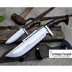 May include: Two knives with wooden handles and leather sheaths. The larger knife has a long, curved blade and a silver finish. The smaller knife has a straight blade and a brown finish. The knives are on a wooden surface with a hammer and a pair of pliers in the background. The text "Heritage Forged" is visible in the lower right corner of the image.  Forged with Passion, Built for Legends.