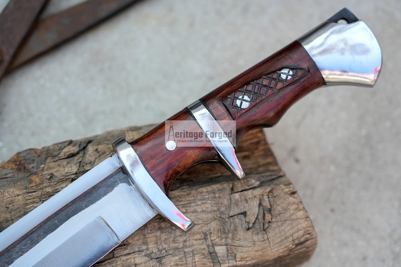May include: A close-up of a silver knife with a wooden handle. The handle is a dark red color and has a decorative pattern. The knife is resting on a piece of wood. The text "Heritage Forged" is visible on the handle.