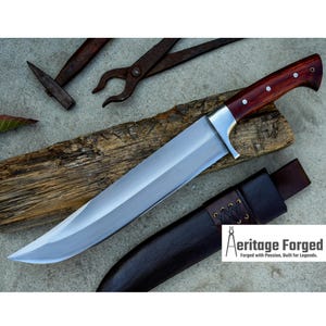 May include: A long, silver blade with a red wooden handle and a leather sheath. The blade is sharp and curved, and the handle is decorated with brass accents. The sheath is brown leather with a braided strap. The text "Heritage Forged" is visible in the image. "Forged with Passion, Built for Legends."