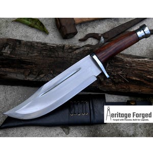 May include: A large, polished steel knife with a dark wood handle and a leather sheath. The blade is wide and curved, with a silver bolster. The knife is resting on a wooden surface. The text "Aeritage Forged" is visible.