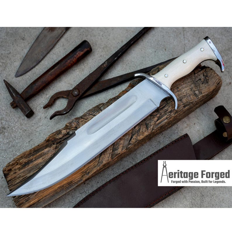 Op de afbeelding: Een groot, zilverkleurig mes met een witte handgreep en een bruine leren schede. Het mes ligt op een stuk hout. De tekst "Heritage Forged" is zichtbaar in de rechterbenedenhoek van de afbeelding. "Forged with Passion, Built for Legends."