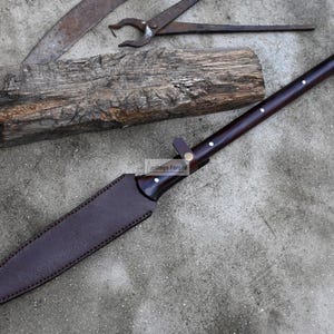 May include: A brown leather-wrapped spear with a silver tip and a wooden handle. The spear is lying on a concrete surface with a piece of wood and a metal tool in the background.