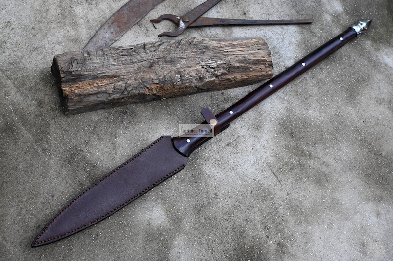 May include: A brown leather-wrapped spear with a silver tip and a wooden handle. The spear is lying on a concrete surface with a piece of wood and a metal tool in the background.