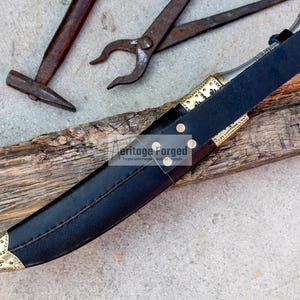 May include: A black leather scabbard with a gold-trimmed dagger inside. The scabbard has a decorative brass buckle and the dagger has a gold-colored handle and blade. The scabbard and dagger are lying on a piece of wood.