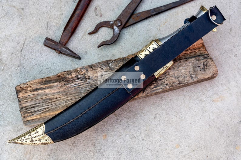 May include: A black leather scabbard with a gold-trimmed dagger inside. The scabbard has a decorative brass buckle and the dagger has a gold-colored handle and blade. The scabbard and dagger are lying on a piece of wood.