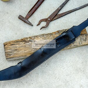 May include: A black leather scabbard with a curved blade. The scabbard has a metal tip and a metal buckle. The scabbard is lying on a piece of wood. The text "Heritage Forged" is visible on the scabbard.