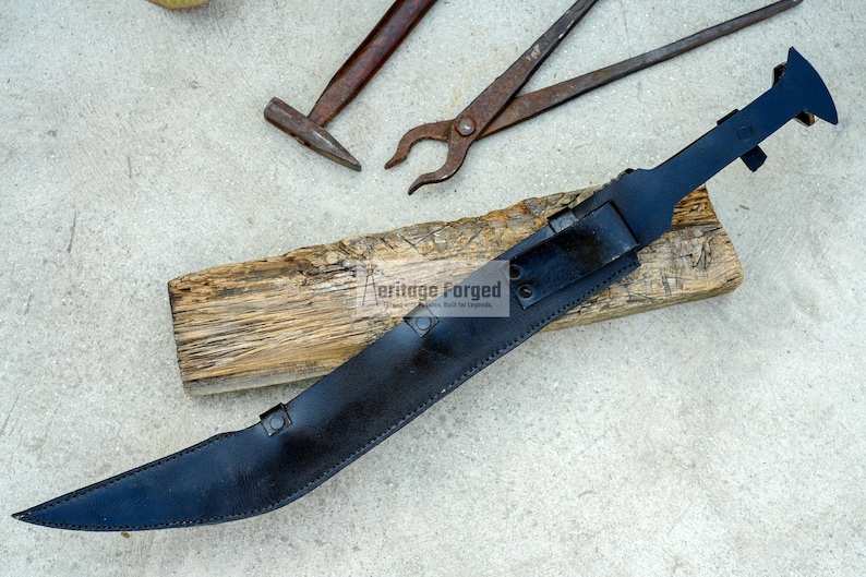 May include: A black leather scabbard with a curved blade. The scabbard has a metal tip and a metal buckle. The scabbard is lying on a piece of wood. The text "Heritage Forged" is visible on the scabbard.
