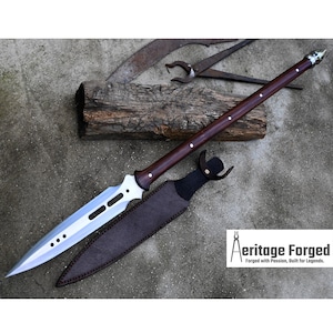 May include: A silver and brown spear with a wooden handle and a leather sheath. The spear has a pointed tip and a serrated blade. The sheath is brown leather with a silver buckle. The spear is on a wooden surface. The text "Heritage Forged, Forged with Passion, Built for Legends." is in the lower right corner of the image.