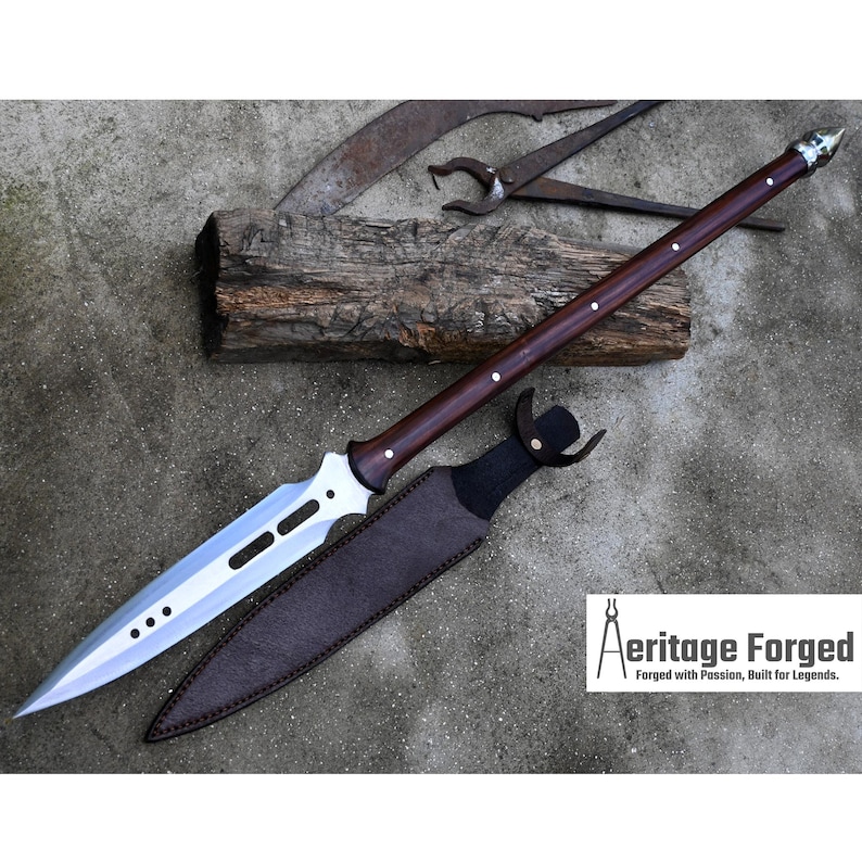 May include: A silver and brown spear with a wooden handle and a leather sheath. The spear has a pointed tip and a serrated blade. The sheath is brown leather with a silver buckle. The spear is on a wooden surface. The text "Heritage Forged, Forged with Passion, Built for Legends." is in the lower right corner of the image.