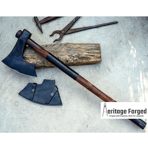 Op de afbeelding: Een zwarte bijl met een houten steel en een zwarte leren schede. De bijl heeft een gebogen blad en een afgeronde kop. De steel is omwikkeld met zwarte tape. De schede heeft een drukknoop sluiting. De bijl ligt op een stuk hout. De tekst "Heritage Forged" is zichtbaar in de rechter benedenhoek van de afbeelding.  Gesmeed met passie, gebouwd voor legendes.