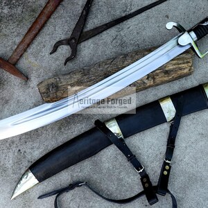 May include: A large, curved sword with a silver blade and black handle, resting on a wooden plank. The sword is accompanied by a black sheath with gold accents and leather straps. Other tools are in the background.